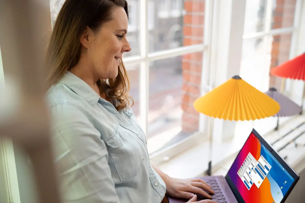 Persona sonriendo mientras teclea en un portátil junto a una ventana, con lámparas de diseño de colores al fondo